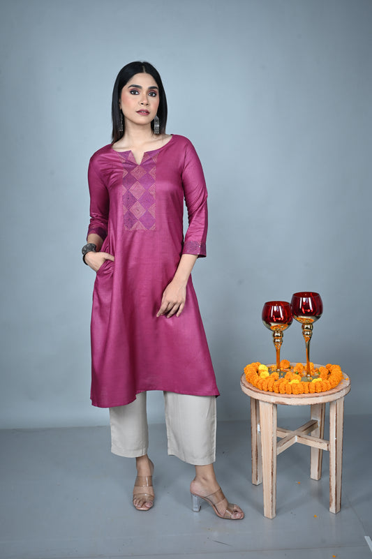 Woman wearing a purple traditional outfit with white pants, standing next to a small wooden stool with decorative items on a gray background.