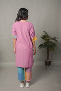 Woman wearing a pink kurta with colorful pants standing in a minimalistic room with a plant.