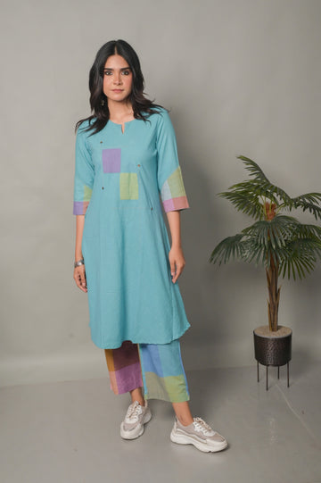 Woman wearing a light blue dress with colorful accents standing next to a potted plant on a plain background