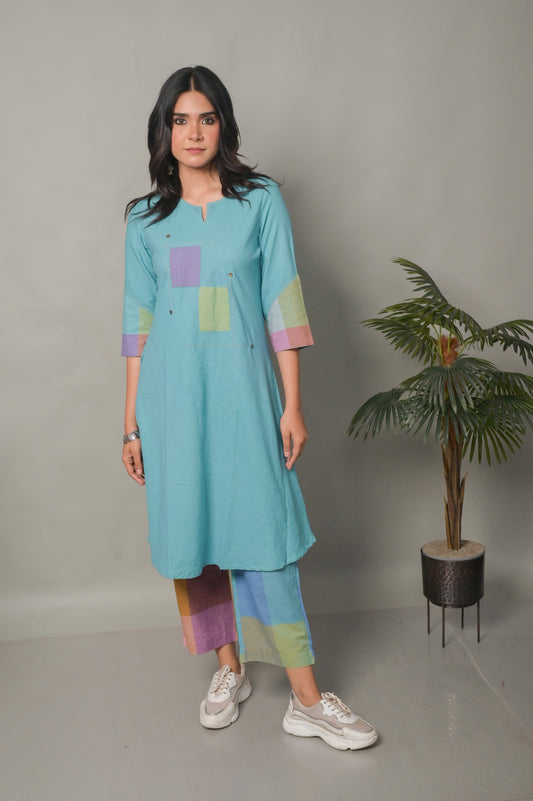 Woman wearing a light blue dress with colorful accents standing next to a potted plant on a plain background