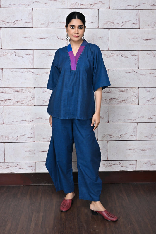 A person wearing a relaxed fit, handwoven top in blue color with a V-neckline and a matching blue pants, standing against a white brick wall backdrop.