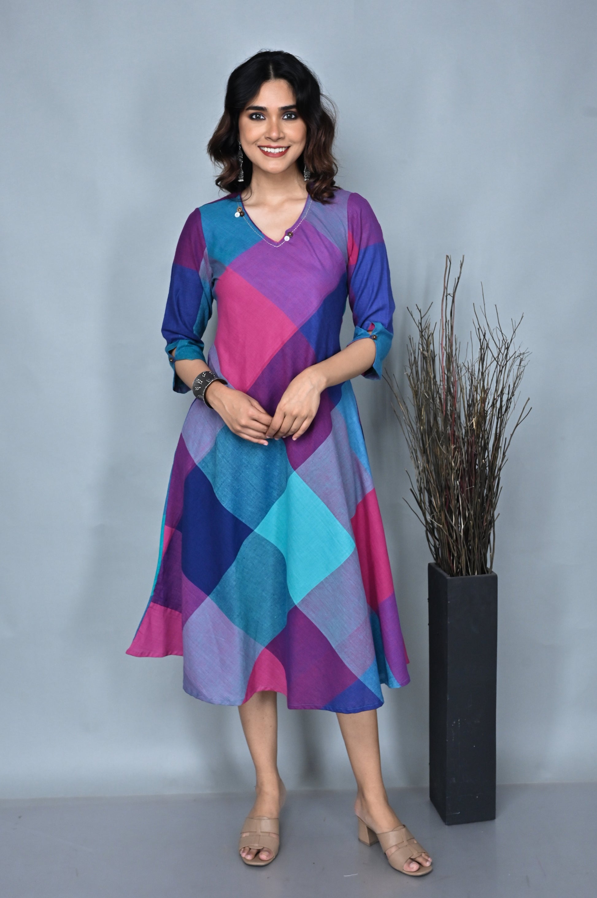 A woman standing against a grey background, wearing a pink and green handwoven checkered dress with kantha embroidery detailing on the neck.