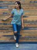 A woman posing with one hand against a wooden background, wearing a blue and green floral printed cotton short top and blue jeans.