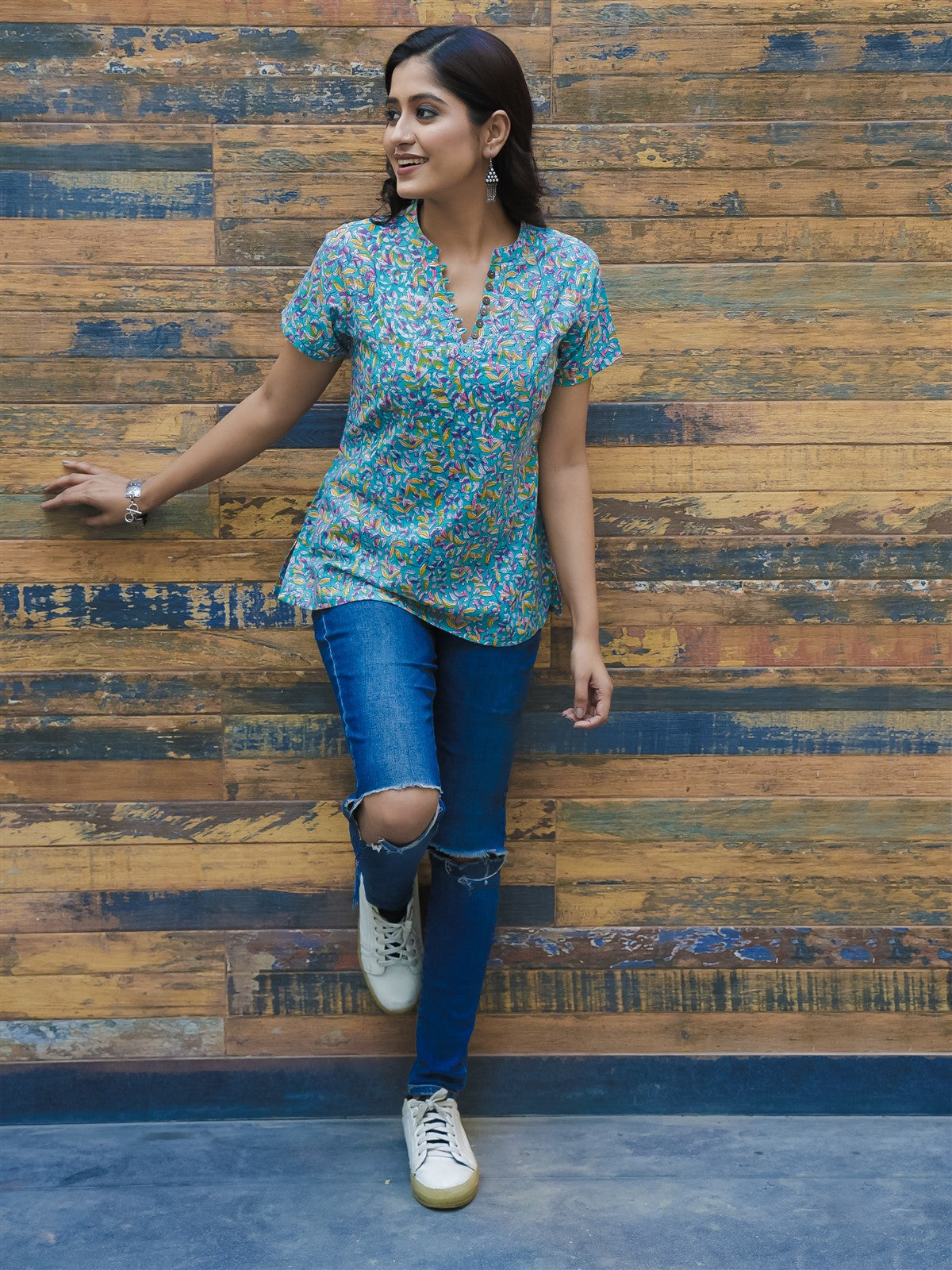A woman posing with one hand against a wooden background, wearing a blue and green floral printed cotton short top and blue jeans.