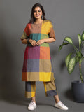 A woman wearing a multicolor checkered handwoven top with elbow-length sleeves and matching trousers, standing against a gray background with a potted plant beside her.