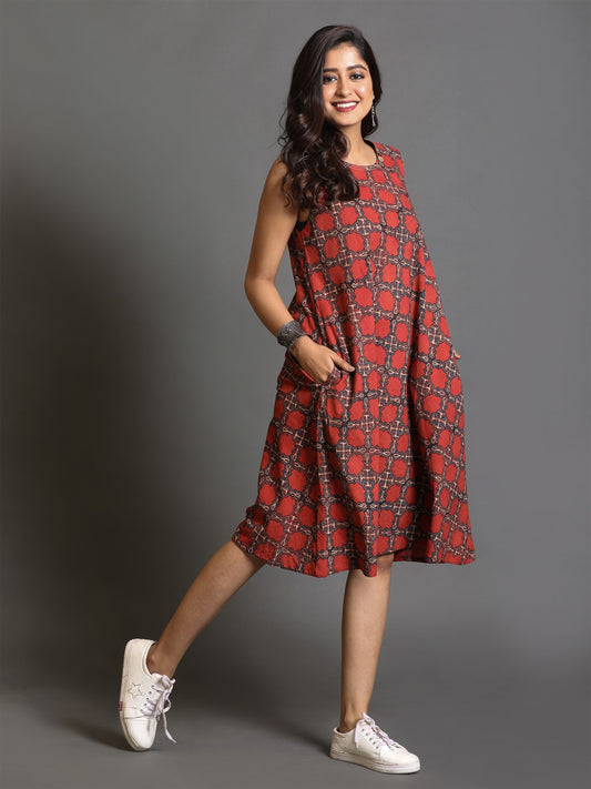 A woman standing, wearing a red ajrakh block-printed sleeveless dress with kantha hand embroidery detailing, paired with white sneakers.