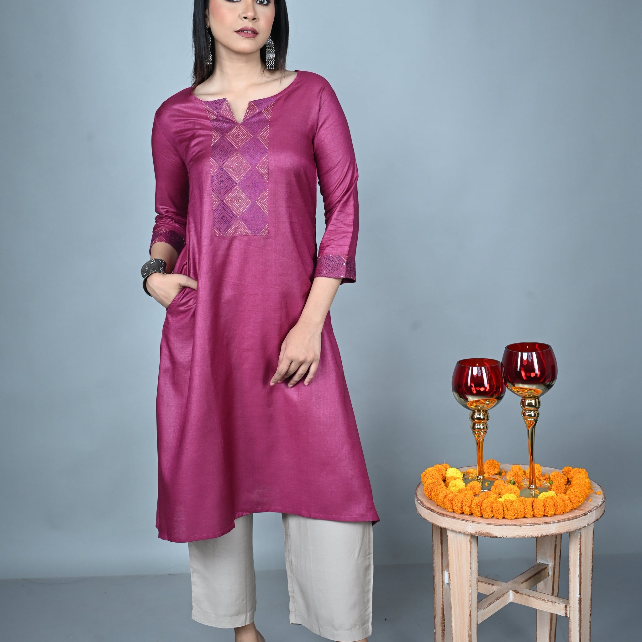 Woman wearing a purple traditional outfit with white pants, standing next to a small wooden stool with decorative items on a gray background.