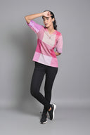 A person wearing a pink and purple checks hand woven top with Kantha hand embroidery detailing, paired with black pants and white sneakers, standing in front of a grey background with a potted plant in the background.