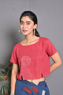 A woman wearing a red handwoven crop top with geometric hand embroidery detailing, paired with blue denim shorts.