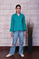 A woman wearing an emerald green checkered high low hand embroidered top with button-up front, paired with blue ripped jeans.