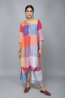 A woman standing against a brick wall backdrop wearing a multicolored, handwoven, cotton kurta with buttons on slits, paired with light-colored trousers and red closed-toe shoes.