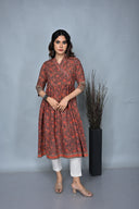 A woman standing against a grey background, wearing a rust-colored cotton hand block printed Ajrakh A-line dress with hand embroidery detailing on the neck, paired with white pants.
