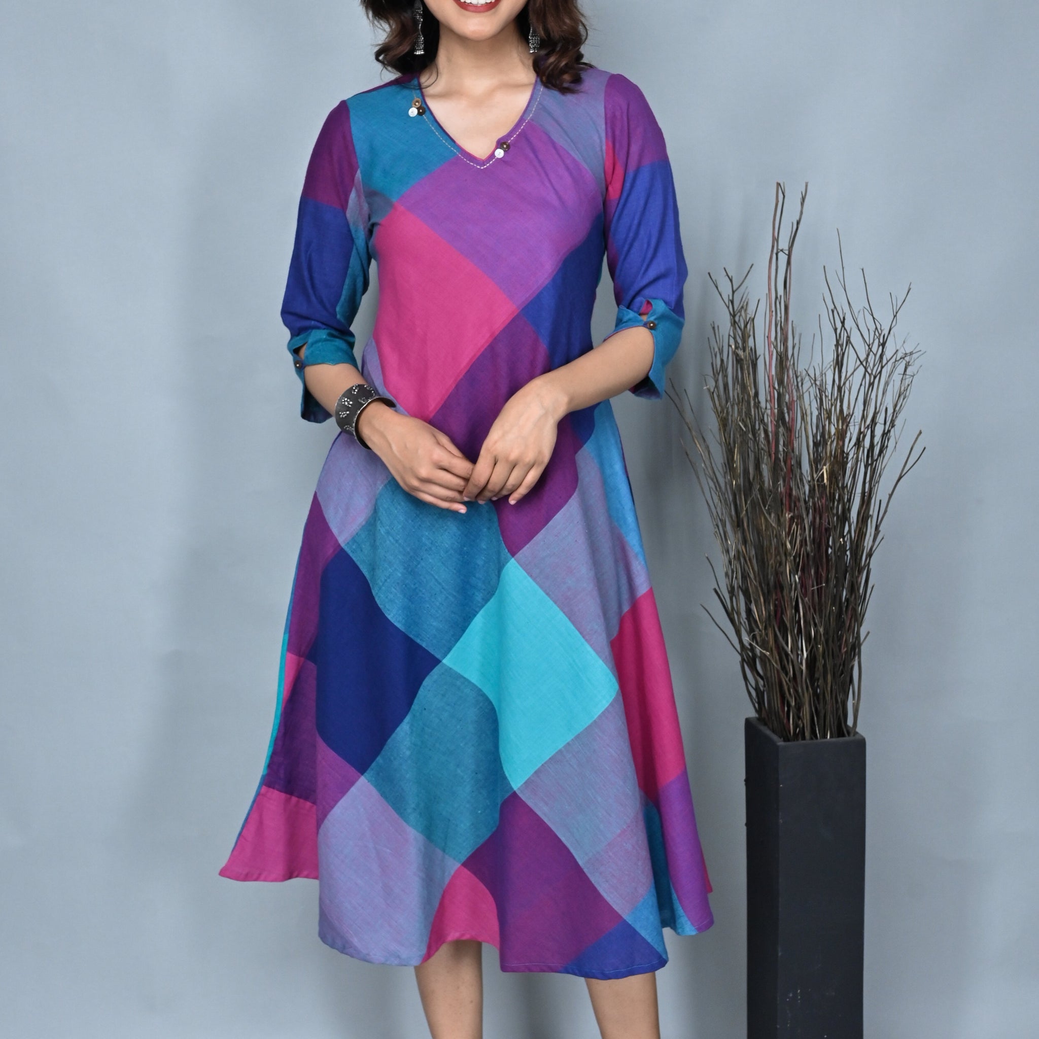 A woman standing against a grey background, wearing a pink and green handwoven checkered dress with kantha embroidery detailing on the neck.