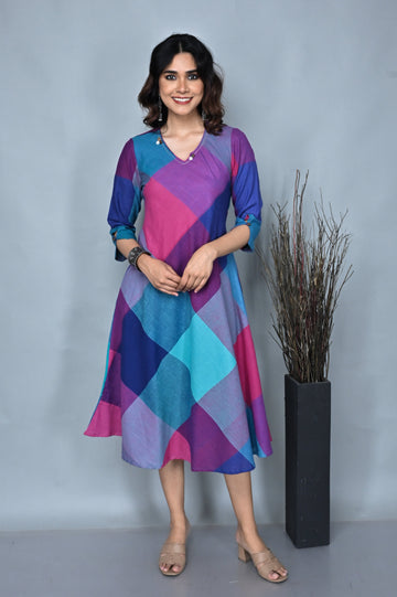 A woman standing against a grey background, wearing a pink and green handwoven checkered dress with kantha embroidery detailing on the neck.