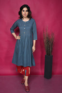 A woman standing against a pink background, wearing a grey pleated kurta with kantha hand embroidery, paired with red pants and matching earrings.