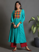 A woman wearing a green handwoven pleated kurta with a multi-color yoke and mirror work, standing in a room with a potted plant and red-colored floor cushion.
